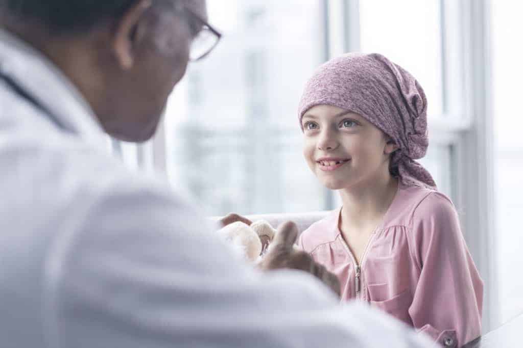 A girl with leukemia sits near a window while meeting with her doctor. The patient is wearing a bandana. She is smiling up at her doctor as he gives her good news.
