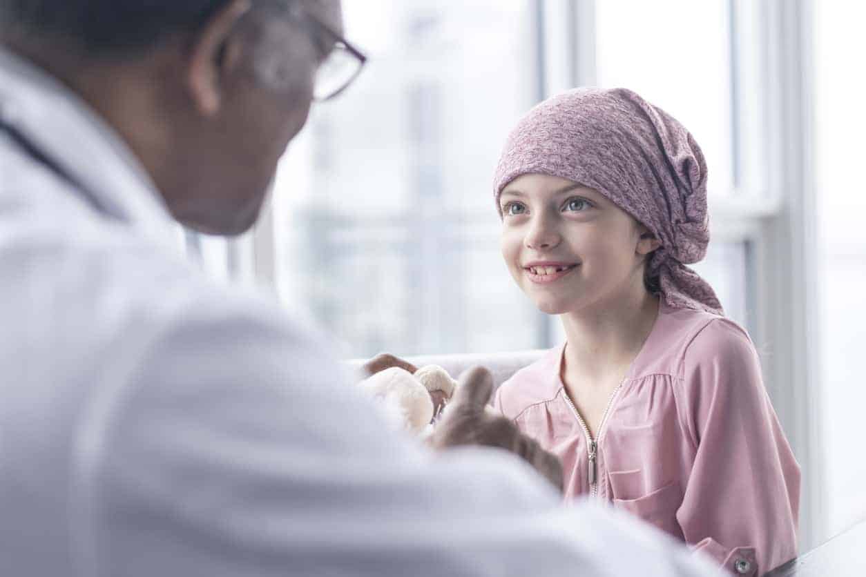 A girl with leukemia sits near a window while meeting with her doctor. The patient is wearing a bandana. She is smiling up at her doctor as he gives her good news.